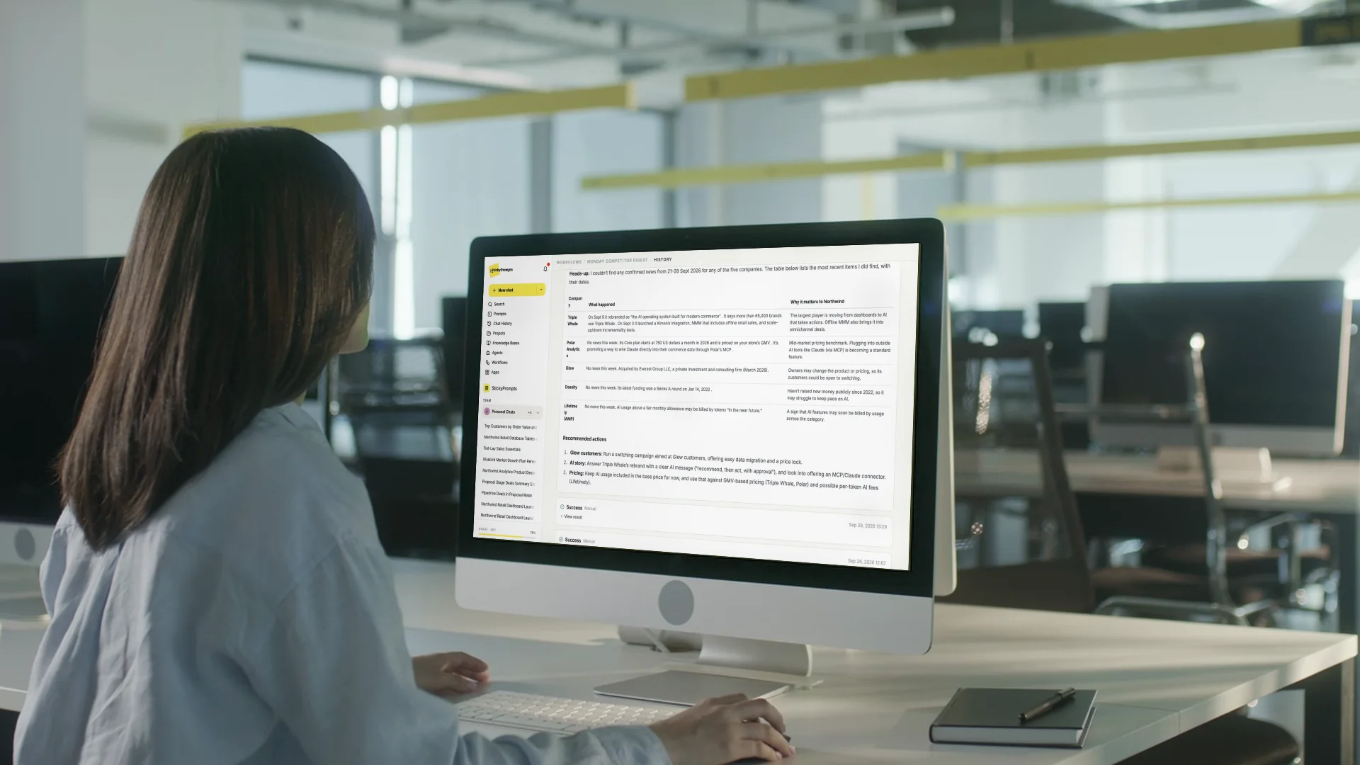 A woman at an iMac in a large open-plan office, a StickyPrompts workflow result on screen